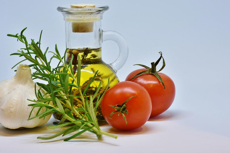 A glass flask of olive oil next to tomatoes, garlic, and herbs