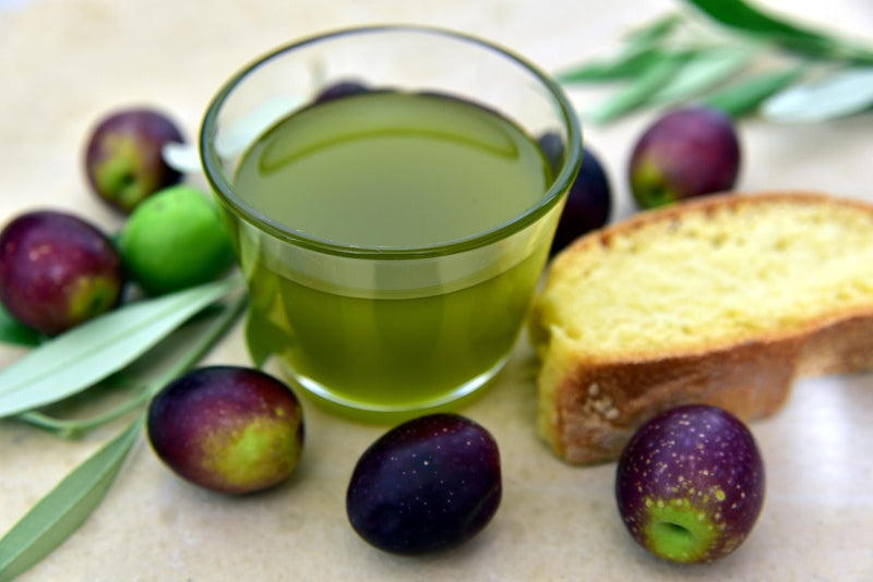 A cup of olive oil next to some olives and bread on a table